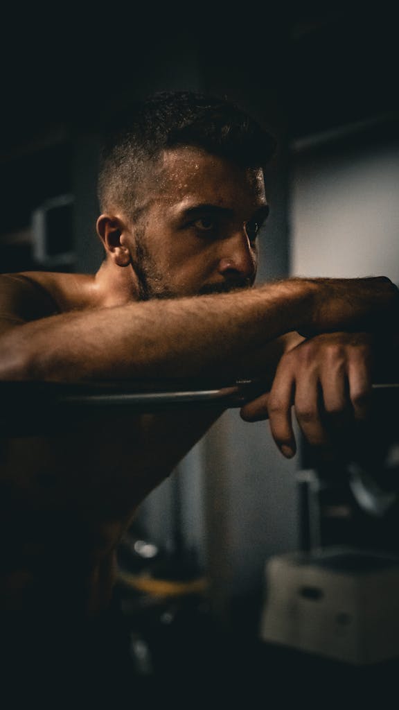 A focused male athlete leaning on a barbell, deep in thought, inside a dimly lit gym.